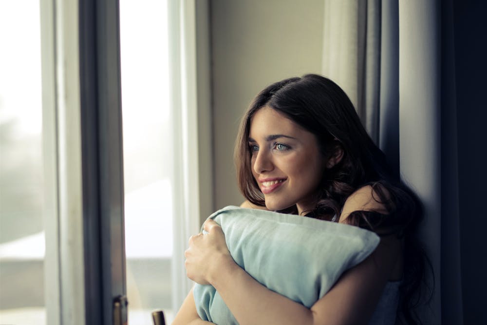 Brown-haired woman sitting next to a glass window and hugging a pillow.